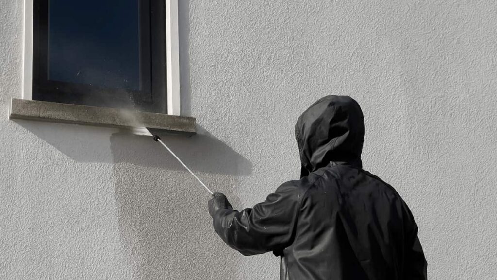 Vakman in zwarte regenjas die een crepi gevel rond een raam reinigt met een hogedrukspuit.